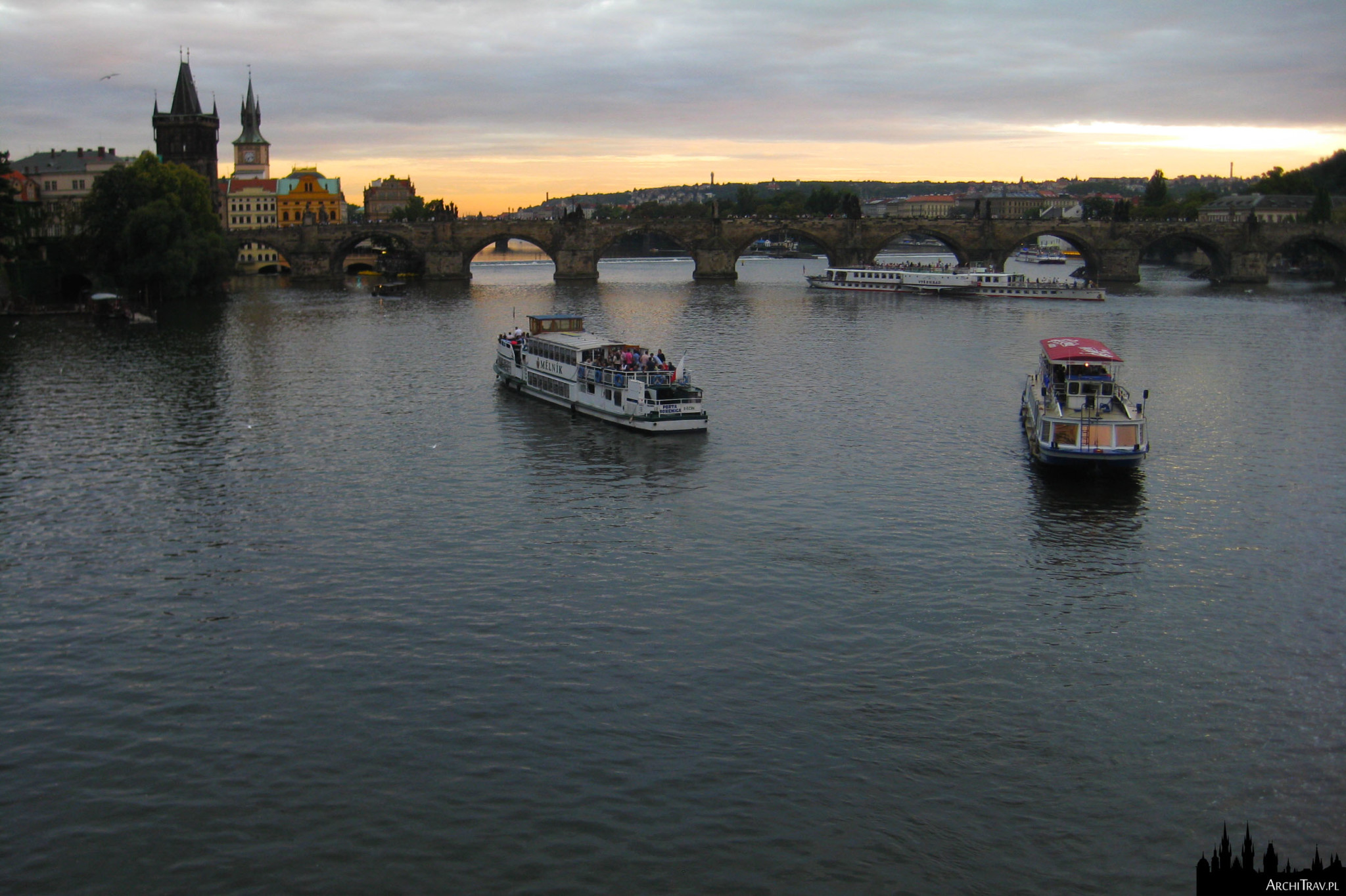 widok na Wełtawę, płynie kilka stateczków, w oddali Most Karola, zachód słońca
