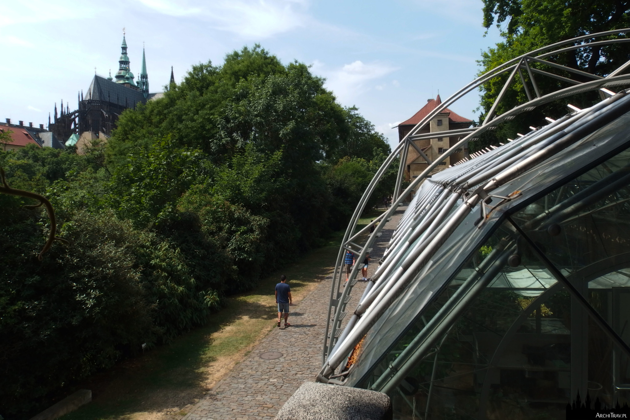 po prawej na pierwszym planie kawałek Oranżerii (Ogrody Królewskie w Pradze), nowoczesnego budynku ze stalowymi i szklanymi elementami, w dole ścieżka, obok niej bujna zieleń, w oddali strzelista sylweta katedry św. Wita